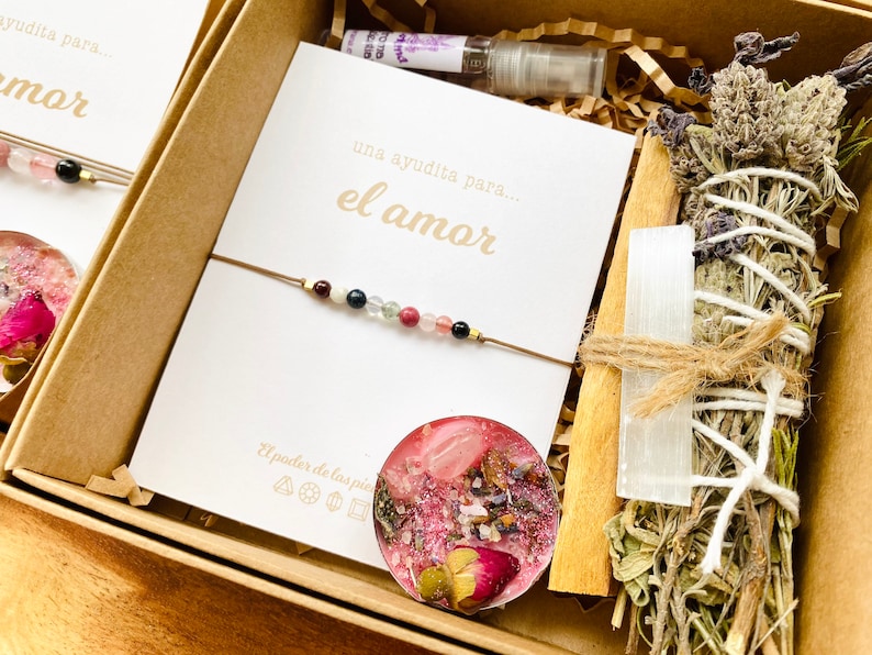 Op de afbeelding: Een cadeauset voor de liefde met een kaart waarop staat "una ayudita para... el amor" (een klein beetje hulp voor... de liefde), een armband met gekleurde kralen, een kaars met roze was en gedroogde bloemen, een wierookstokje met gedroogde kruiden en een witte kristal, en een houten stok.
