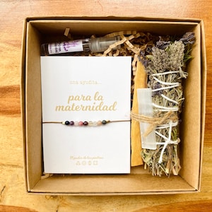 May include: A gift box containing a bracelet with colorful beads, a card with Spanish text, a bundle of dried herbs, a crystal, a stick of wood, and a small spray bottle. The box is brown and sits on a wooden surface.