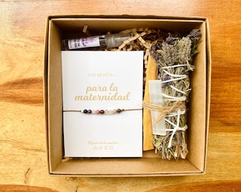 May include: A gift box containing a bracelet with colorful beads, a card with Spanish text, a bundle of dried herbs, a crystal, a stick of wood, and a small spray bottle. The box is brown and sits on a wooden surface.