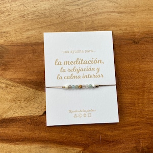 Op de afbeelding: Een witte kaart met de tekst "una ayudita para... la meditaci&oacute;n, la relajaci&oacute;n y la calma interior" in gouden letters. Een bruin koordarmband met witte en groene kralen is om de kaart gebonden.