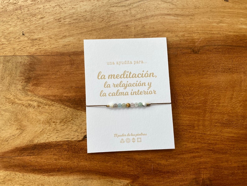 Op de afbeelding: Een witte kaart met de tekst "una ayudita para... la meditaci&oacute;n, la relajaci&oacute;n y la calma interior" in gouden letters. Een bruin koordarmband met witte en groene kralen is om de kaart gebonden.