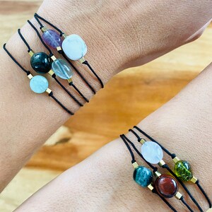 Può includere: Diversi braccialetti con pietre preziose con cordoncini neri e dettagli metallici color oro sono esposti sui polsi. I braccialetti presentano varie pietre colorate, tra cui viola, verde, blu e bianco, di forma rotonda e ovale.