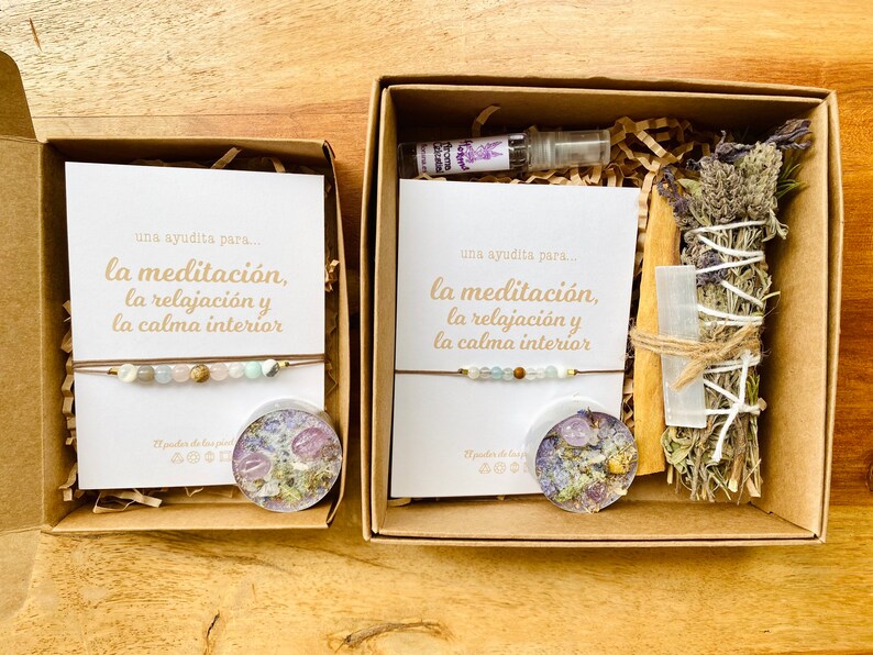 Op de afbeelding: Twee bruine cadeaudozen met open deksels. De dozen bevatten items voor meditatie en ontspanning. De linker doos bevat een armband, een kaars en een kaart met de tekst "una ayudita para... la meditaci&oacute;n, la relajaci&oacute;n y la calma interior". De rechter doos bevat een wierookstokje, een kristal, een flesje etherische olie en een kaart met de tekst "una ayudita para... la meditaci&oacute;n, la relajaci&oacute;n y la calma interior".