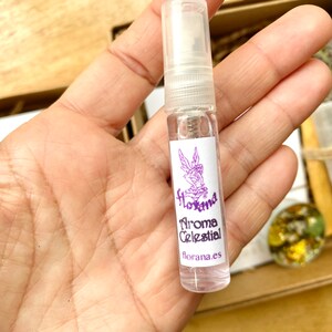 May include: A clear glass spray bottle with a white label featuring the text "Florana Aroma Celestial" and a purple fairy illustration. The bottle is held in a hand, with other items in a wooden box visible in the background.