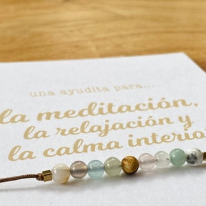Op de afbeelding: Een bruin koordarmband met een verscheidenheid aan gekleurde kralen, waaronder wit, grijs, lichtblauw, groen en bruin. De armband ligt op een witte kaart met de tekst "una ayudita para... la meditaci&oacute;n, la relajaci&oacute;n y la calma interior" in gouden letters.