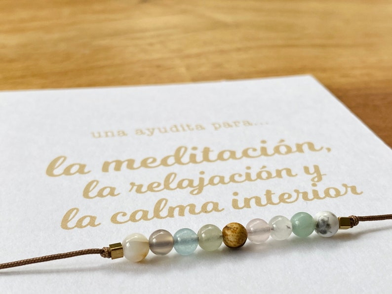 Op de afbeelding: Een bruin koordarmband met een verscheidenheid aan gekleurde kralen, waaronder wit, grijs, lichtblauw, groen en bruin. De armband ligt op een witte kaart met de tekst "una ayudita para... la meditaci&oacute;n, la relajaci&oacute;n y la calma interior" in gouden letters.