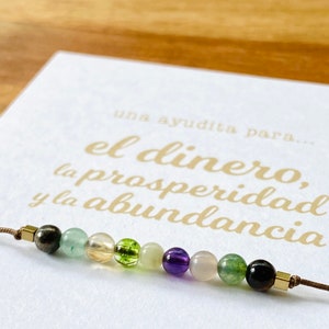 May include: A beaded bracelet featuring a variety of colorful gemstones, including green, yellow, purple, and black beads. The bracelet is strung on a brown cord and is displayed on a white card with gold text that reads "el dinero, la prosperidad y la abundancia."