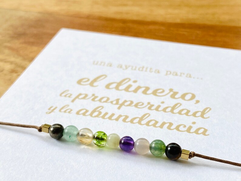 May include: A beaded bracelet featuring a variety of colorful gemstones, including green, yellow, purple, and black beads. The bracelet is strung on a brown cord and is displayed on a white card with gold text that reads "el dinero, la prosperidad y la abundancia."