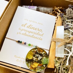 May include: A gift box containing a card with gold text reading "el dinero, la prosperidad y la abundancia", a beaded bracelet, a bundle of dried herbs, a selenite wand, and a small bottle. The box is filled with brown paper.