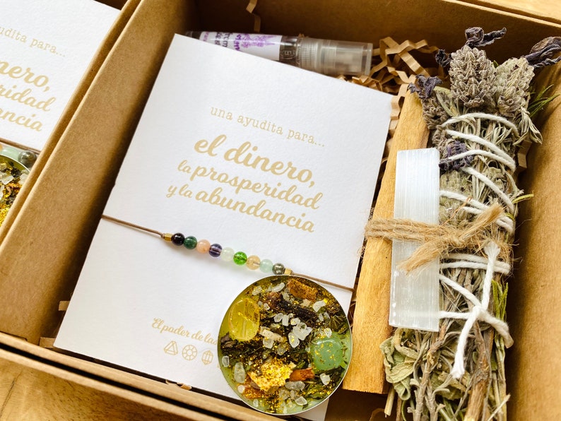 May include: A gift box containing a card with gold text reading "el dinero, la prosperidad y la abundancia", a beaded bracelet, a bundle of dried herbs, a selenite wand, and a small bottle. The box is filled with brown paper.