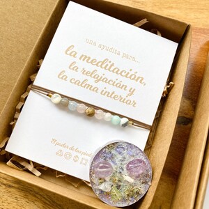 Op de afbeelding: Een bruine kartonnen doos met een witte kaart erin. De kaart heeft Spaanse tekst die luidt "una ayudita para... la meditaci&oacute;n, la relajaci&oacute;n y la calma interior". De doos bevat ook een armband met kralen en een kleine kaars met gedroogde bloemen en kristallen.