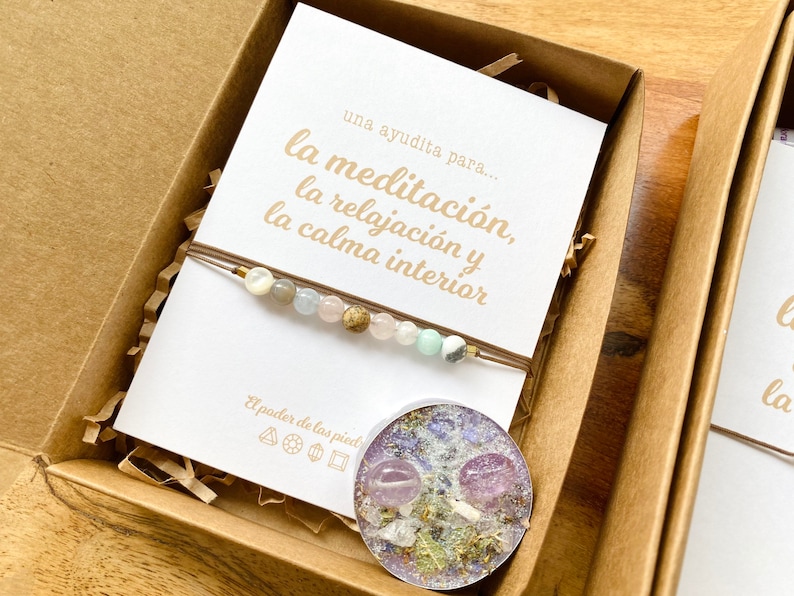 Op de afbeelding: Een bruine kartonnen doos met een witte kaart erin. De kaart heeft Spaanse tekst die luidt "una ayudita para... la meditaci&oacute;n, la relajaci&oacute;n y la calma interior". De doos bevat ook een armband met kralen en een kleine kaars met gedroogde bloemen en kristallen.