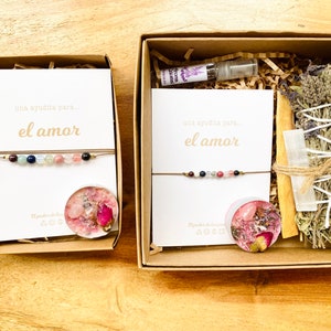 Op de afbeelding: Twee cadeaudozen met bruine deksels en witte kaartjes waarop staat "una ayudita para... el amor" (een klein beetje hulp voor... de liefde). De dozen bevatten een armband, een kaars, een wierookstokje en een flesje etherische olie.