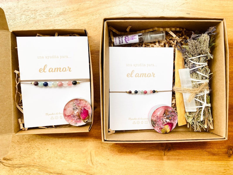 Op de afbeelding: Twee cadeaudozen met bruine deksels en witte kaartjes waarop staat "una ayudita para... el amor" (een klein beetje hulp voor... de liefde). De dozen bevatten een armband, een kaars, een wierookstokje en een flesje etherische olie.