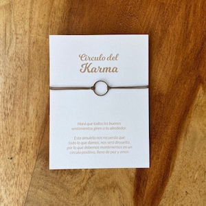 Op de afbeelding: Een minimalistische armband met een kleine, cirkelvormige bedel en een dun, bruin koord. De armband wordt gepresenteerd op een witte kaart met Spaanse tekst, waaronder "Circulo del Karma". De achtergrond is een houten oppervlak.