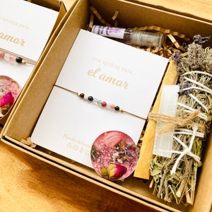 Op de afbeelding: Een bruine doos gevuld met een liefdes-cadeauset. De set bevat een roze-witte kaars, een salie-wierookstokje, een helder kristal, een armband met roze, blauwe en groene kralen en een kaart met de tekst "una ayudita para... el amor".