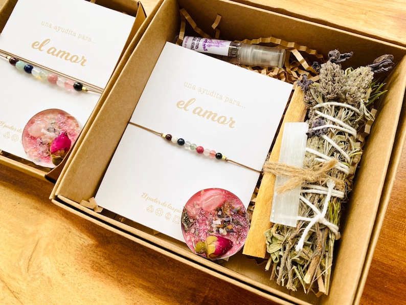 Op de afbeelding: Een bruine doos gevuld met een liefdes-cadeauset. De set bevat een roze-witte kaars, een salie-wierookstokje, een helder kristal, een armband met roze, blauwe en groene kralen en een kaart met de tekst "una ayudita para... el amor".