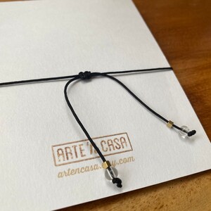 Op de afbeelding: Een zwart koord met een knoop en twee heldere glazen kralen met gouden accenten. Het koord is vastgemaakt aan een witte kaart met de tekst "ARTE Y CASA" en "artencasa.com".