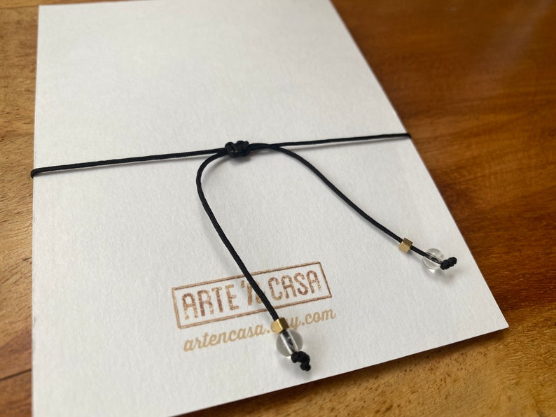 Op de afbeelding: Een zwart koord met een knoop en twee heldere glazen kralen met gouden accenten. Het koord is vastgemaakt aan een witte kaart met de tekst "ARTE Y CASA" en "artencasa.com".