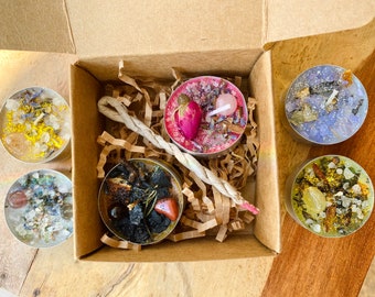May include: A set of five tealight candles in a cardboard box. The candles are filled with various crystals, herbs, and other natural ingredients. The box is filled with brown packing paper.