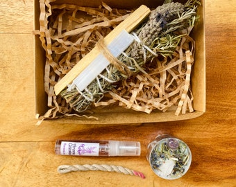 May include: A gift box filled with sage smudge sticks, a palo santo stick, a small bottle of essential oil, a candle with crystals, and a white string. The box is lined with brown paper shreds.