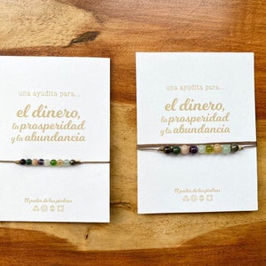 May include: Two beaded bracelets on cards with Spanish text. The bracelets feature a string with colorful gemstones. The cards have text that reads "una ayudita para... el dinero, la prosperidad y la abundancia" and "El poder de las piedras."