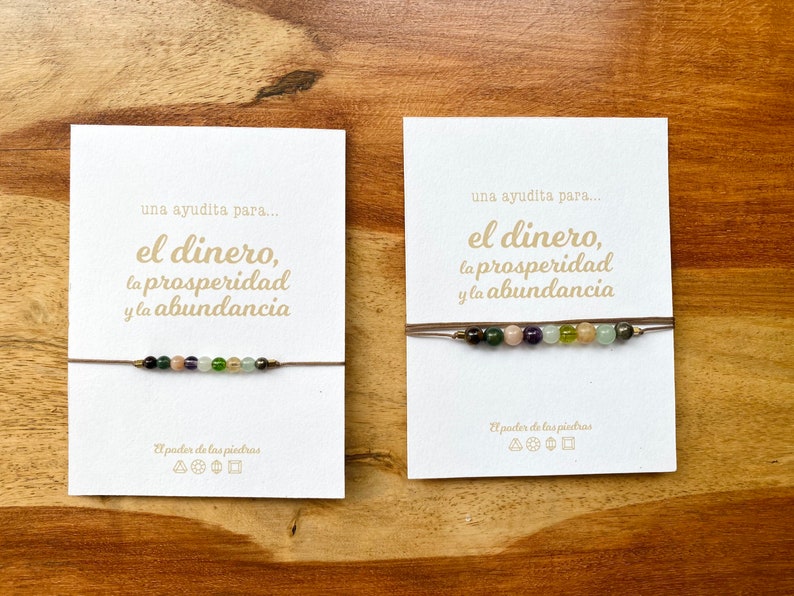 May include: Two beaded bracelets on cards with Spanish text. The bracelets feature a string with colorful gemstones. The cards have text that reads "una ayudita para... el dinero, la prosperidad y la abundancia" and "El poder de las piedras."