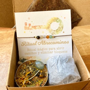 May include: A brown cardboard box contains a ritual kit. The kit includes a card with the text "Ritual Abrecaminos", a bracelet with beads, a candle, and a small package. The candle is decorated with gold glitter and a gemstone.