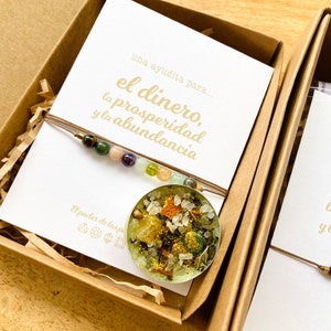 May include: A pendant necklace with a clear, round resin charm filled with colorful stones and dried herbs. The necklace is displayed in a brown box with a card that reads "el dinero, la prosperidad y la abundancia".