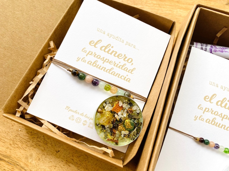 May include: A pendant necklace with a clear, round resin charm filled with colorful stones and dried herbs. The necklace is displayed in a brown box with a card that reads "el dinero, la prosperidad y la abundancia".