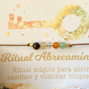 May include: A beaded bracelet with brown cord and gold-colored square accents. The bracelet features a sequence of colorful beads: brown, yellow, clear, orange, and green. The card behind the bracelet has the text "Ritual Abrecaminos".