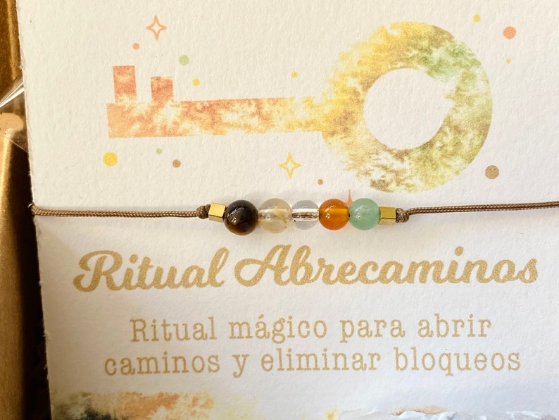 May include: A beaded bracelet with brown cord and gold-colored square accents. The bracelet features a sequence of colorful beads: brown, yellow, clear, orange, and green. The card behind the bracelet has the text "Ritual Abrecaminos".