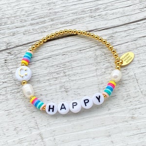 May include: Gold beaded bracelet with white beads, rainbow beads, and white letter beads spelling "HAPPY". The bracelet has a gold charm with the text "Wunder bilden sch&ouml;n".