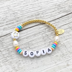 May include: A gold beaded bracelet with white beads, rainbow striped beads, and white letter beads spelling out the name "SOFIA". The bracelet also features a white bead with a gold smiley face.