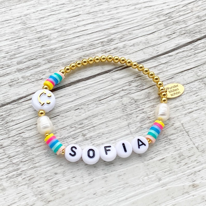 May include: A gold beaded bracelet with white beads, rainbow striped beads, and white letter beads spelling out the name "SOFIA". The bracelet also features a white bead with a gold smiley face.
