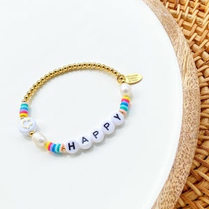 May include: Gold beaded bracelet with white beads that spell out "HAPPY" and rainbow colored beads. The bracelet also has a small gold charm with a smiley face.