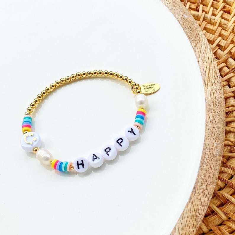 May include: Gold beaded bracelet with white beads that spell out "HAPPY" and rainbow colored beads. The bracelet also has a small gold charm with a smiley face.