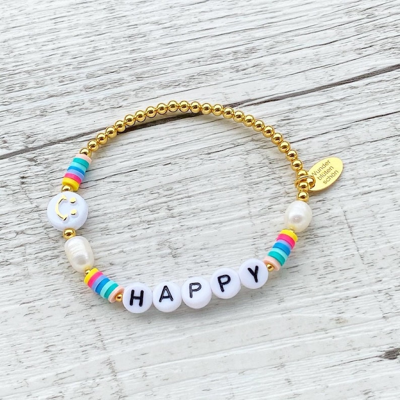 May include: Gold beaded bracelet with white beads, rainbow beads, and white letter beads spelling "HAPPY". The bracelet has a gold charm with the text "Wunder bilden sch&ouml;n".