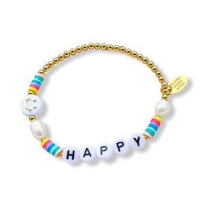 May include: Gold beaded bracelet with white beads spelling out "HAPPY" and a white bead with a smiley face. The bracelet also has a white pearl and a rainbow striped bead. The bracelet has a gold tag that says "Wunder bl&uuml;hen sch&ouml;n".