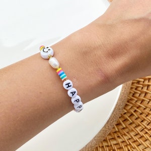 May include: White beaded bracelet with the word "HAPPY" spelled out in black letters, a smiley face bead, and a pearl bead.