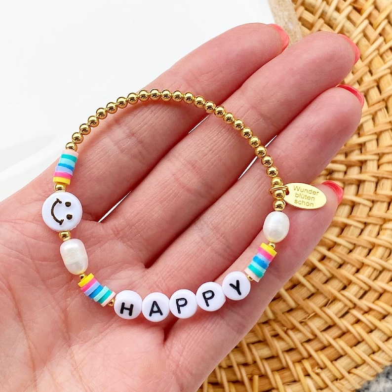 May include: Gold beaded bracelet with white beads, rainbow striped beads, and white letter beads spelling "HAPPY". The bracelet also features a white bead with a smiley face and a gold tag with the text "Wunder bluten sch&ouml;n".
