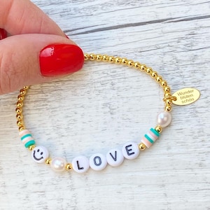 Könnte beinhalten: Goldene Perlenkette mit einer weißen Perle, einem Smiley-Anhänger und dem Wort "LOVE" in weißen Perlen. Das Armband hat auch einen kleinen Anhänger mit dem Text "Wunder bluten schon".