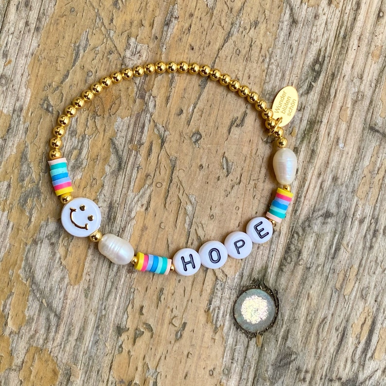 May include: Gold beaded bracelet with white beads, a smiley face charm, and the word "HOPE" spelled out in white letters. The bracelet has a small gold tag that says "Wunder Bl&uuml;hen Sch&ouml;n".