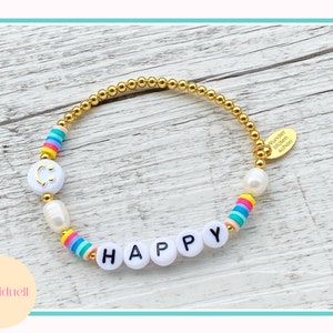 May include: A gold bracelet with white beads and colorful beads that spell out "HAPPY". The bracelet also has a small gold charm with the text "Wunder  bl&uuml;hen  sch&ouml;n".