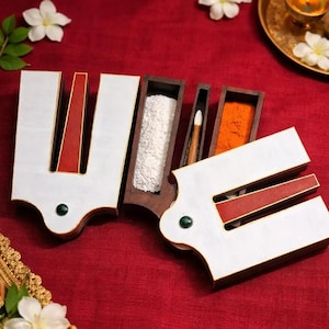 Puede incluir: Un juego de recipientes para tilak de madera en forma de U con superficies pintadas en blanco y rojo y una pequeña incrustación de piedra verde. El juego incluye tres compartimentos estrechos con polvo blanco, un aplicador de madera y polvo naranja.
