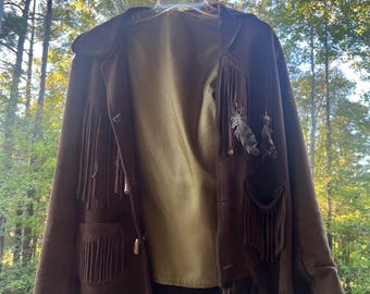 May include: A brown suede jacket with fringe detailing and feather accents. The jacket has a button closure and two pockets.