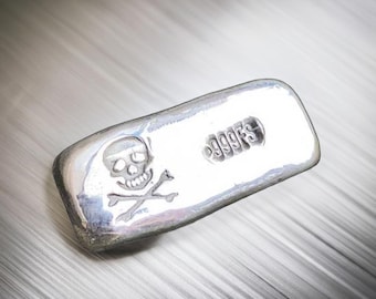Könnte beinhalten: Ein Silberbarren mit einem Totenkopf- und Knochenkreuz-Design und der Aufschrift "999.95" auf der Oberfläche.