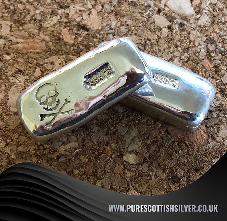 May include: Two silver bars with a skull and crossbones design. The bars are stamped with "999.95" and are on a brown cork surface.