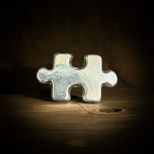 May include: A silver-colored puzzle piece, with a slightly textured surface, sits on a wooden surface. The puzzle piece is the classic two-knob design. The background is a dark, textured fabric.