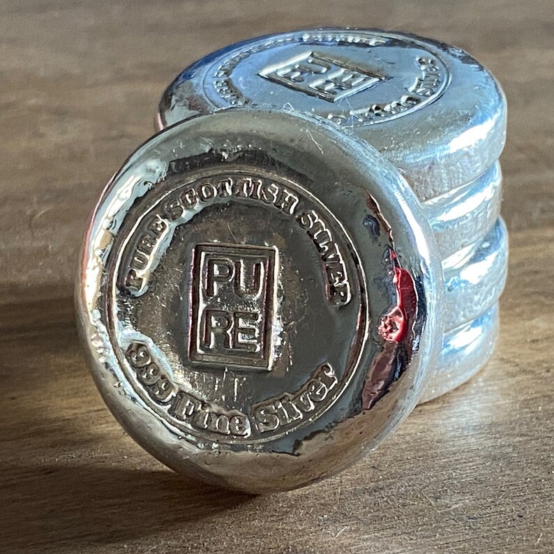 Puede incluir: Una pila de cuatro barras de plata, cada una con las palabras "Pure Scottish Silver 999 Fine Silver" y un logotipo cuadrado que dice "PURE" en may&uacute;sculas.